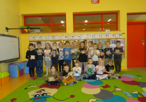 Grupa dzieci pozuje do zdjęcia z przygotowanymi kontrabasami z kartoników, które trzymają w ręku. Dzieci stoi lub siedzi na dywanie.